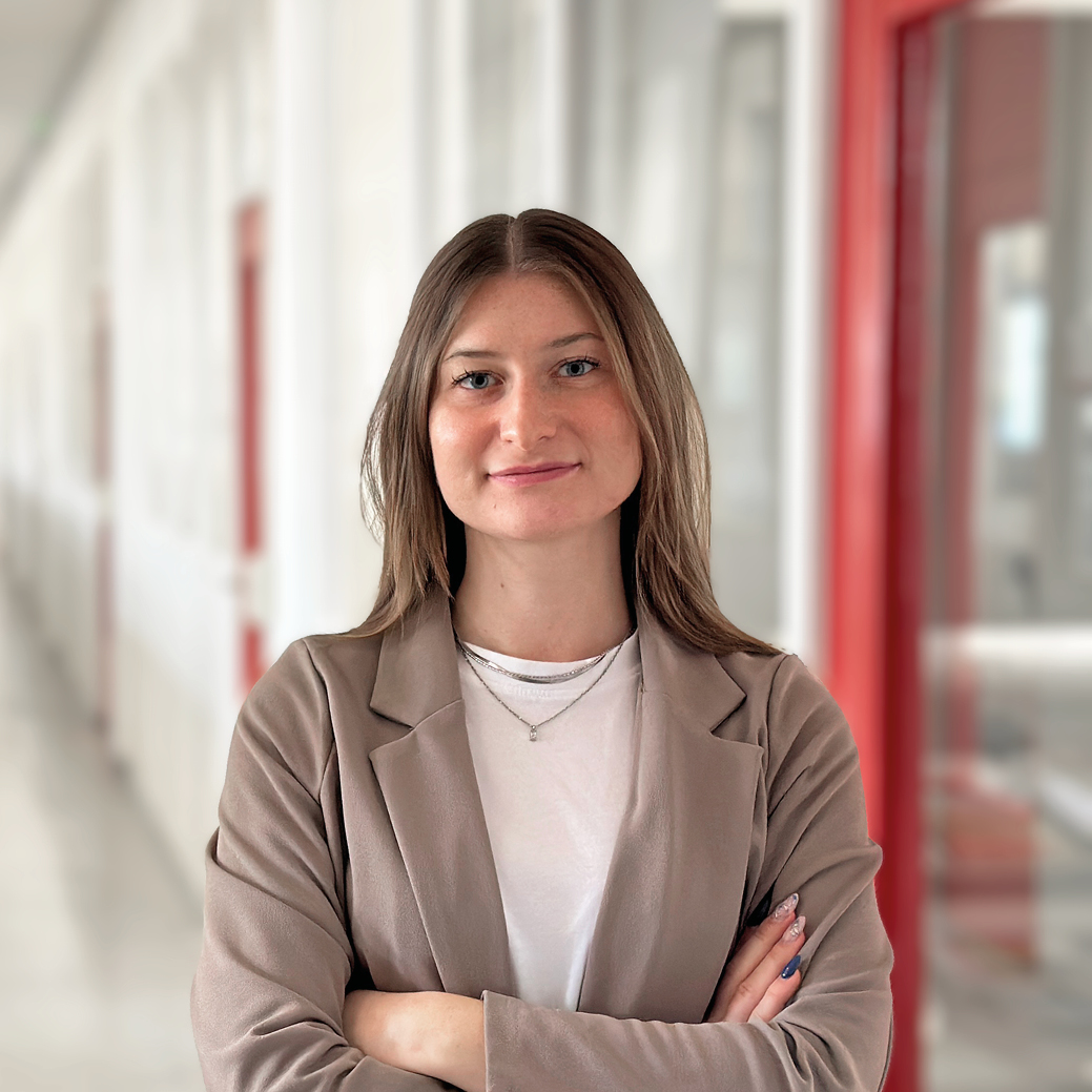 Auf dem Bild ist eine Dame zu sehen, die in die Kamera lächelt. Sie hat braune schulterlange Haare. Sie trägt einen hellbraunen Blazer und darunter ein weißes Oberteil.